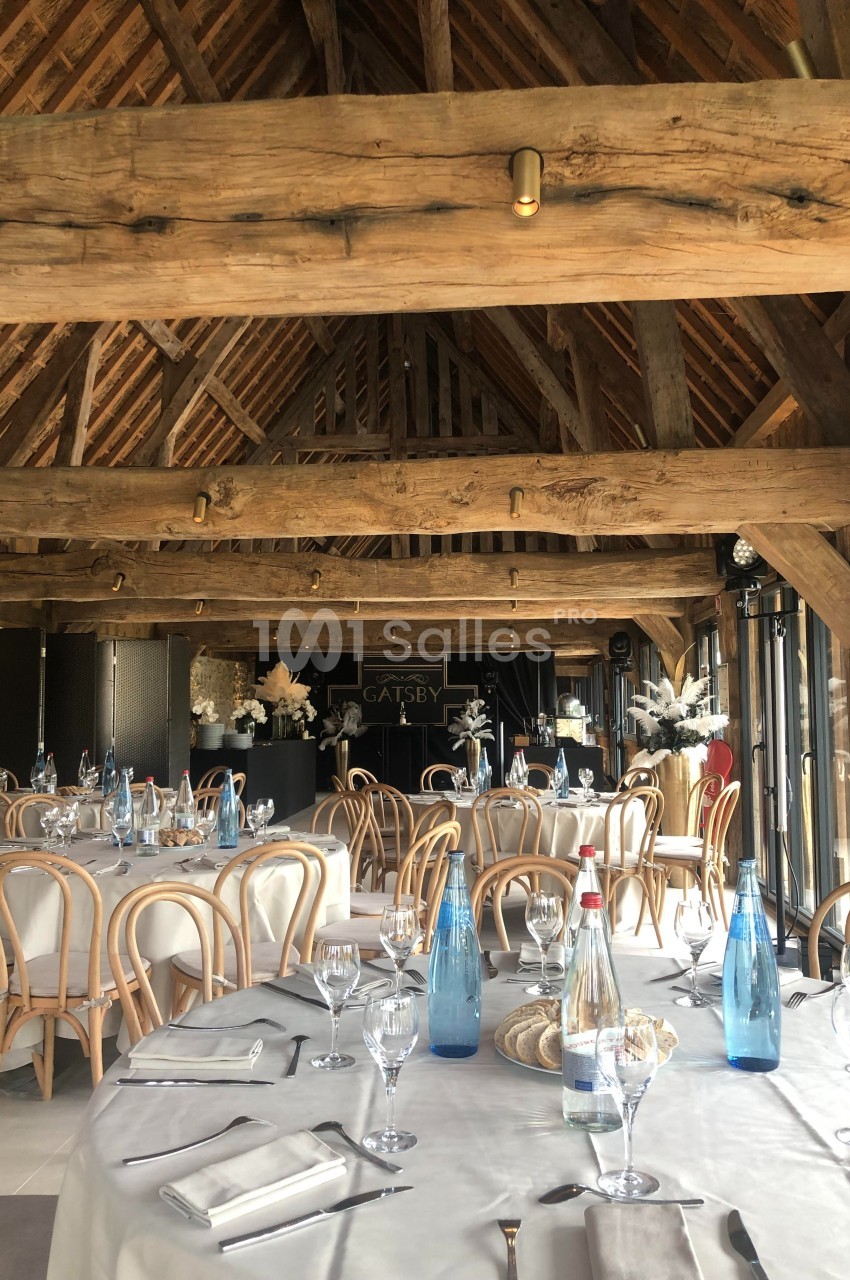 Salle de réception avec charpente en bois apparente, tables rondes dressées avec nappes blanches et bouteilles d'eau.