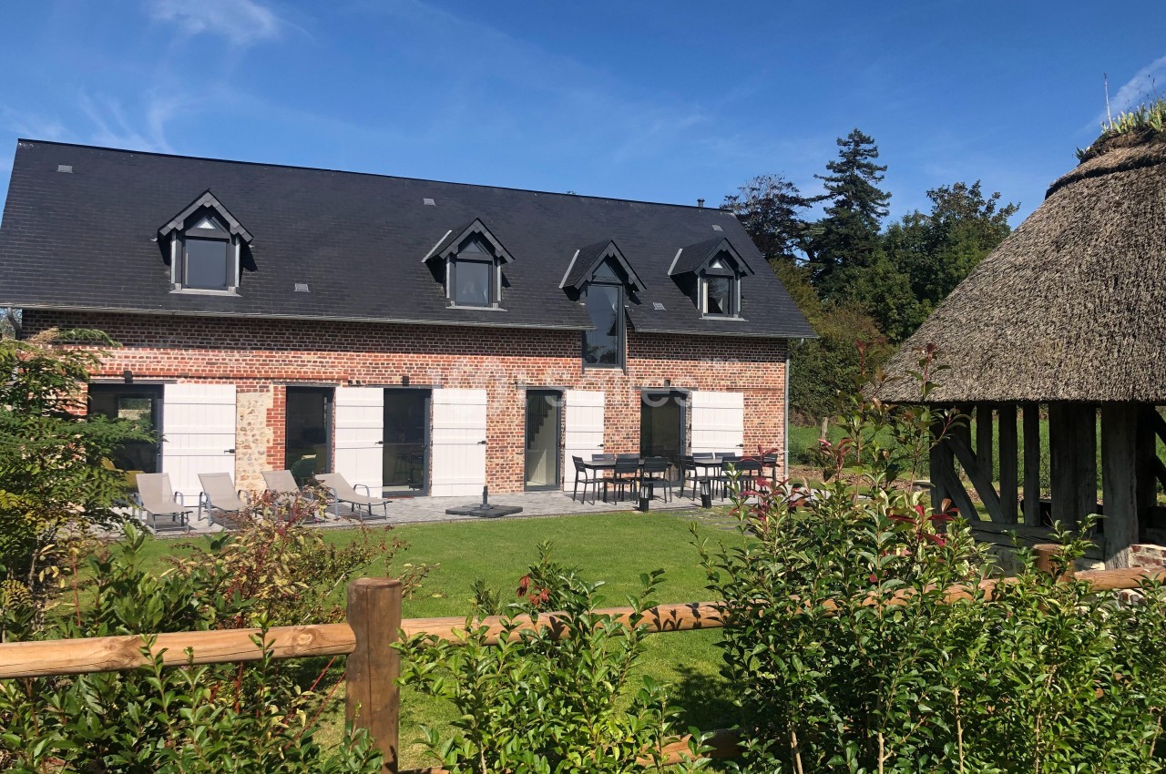Maison en briques avec toit en ardoise, jardin aménagé et terrasse, entourée de verdure et d'une structure en chaume.