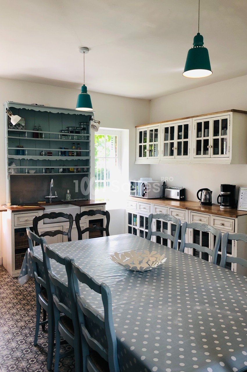 Cuisine lumineuse avec table en bois recouverte d'une nappe à pois, chaises bleues et meubles de rangement rustiques.