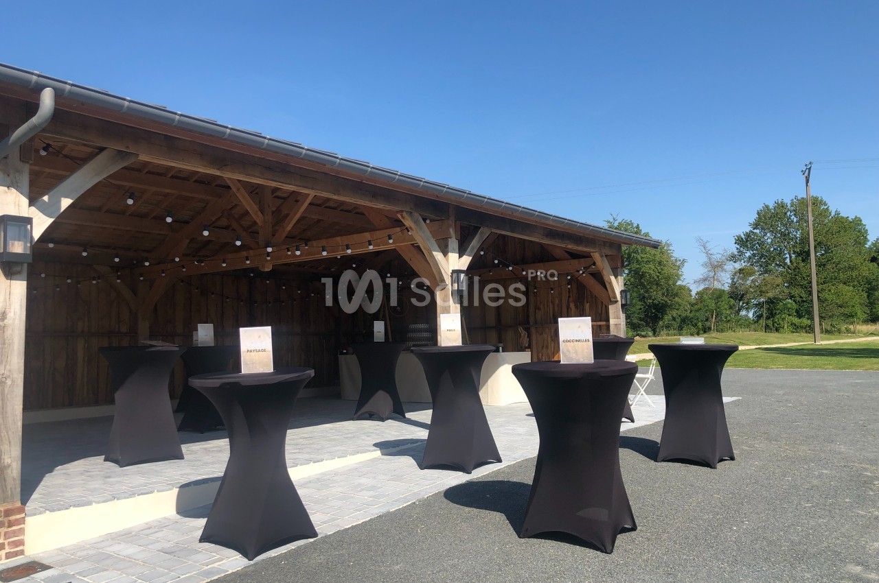 Espace extérieur avec tables hautes noires sous une structure en bois, éclairée par des guirlandes lumineuses.