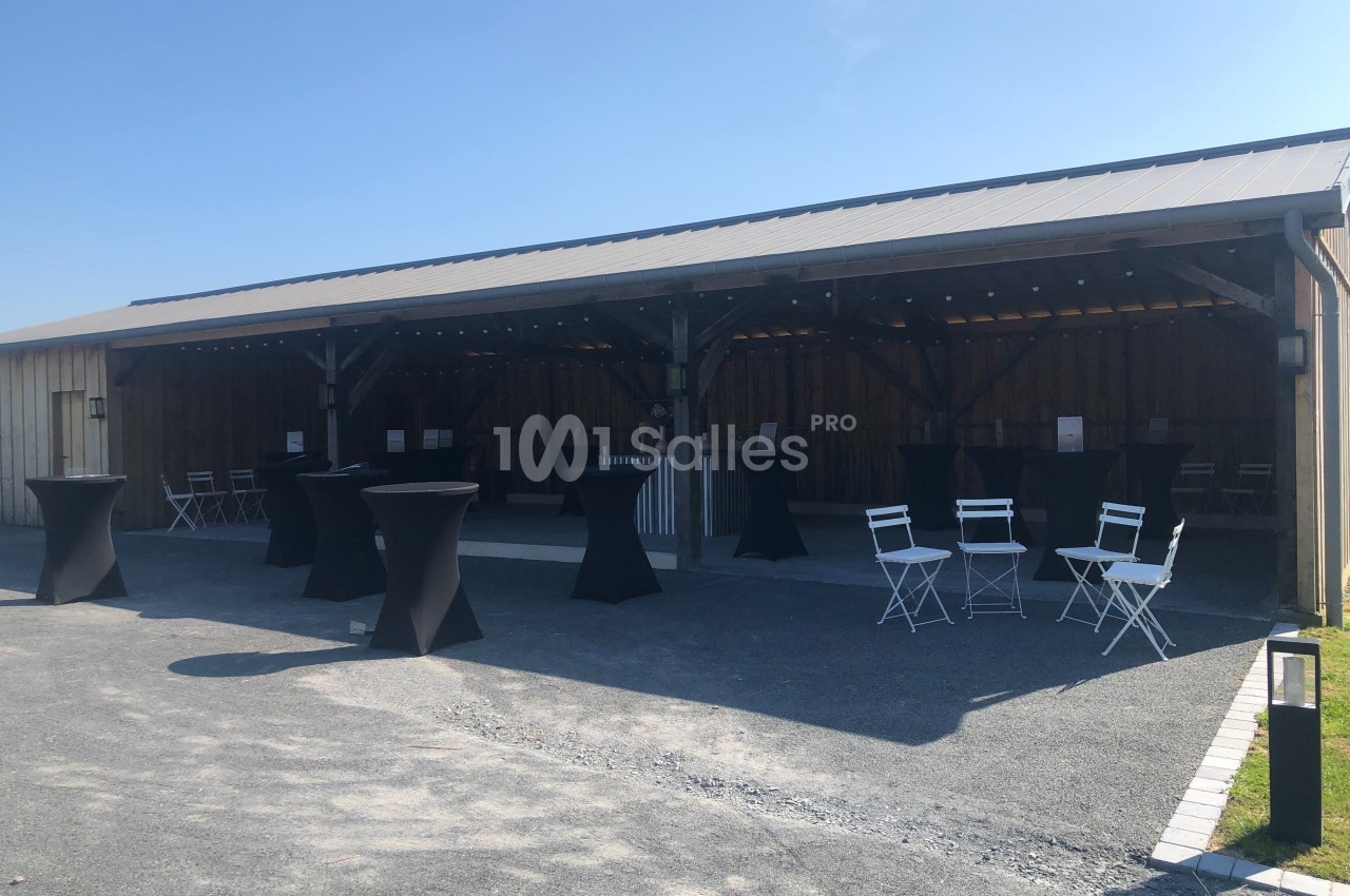 Abri en bois ouvert avec tables hautes noires et chaises blanches disposées sur un sol en gravier, sous un ciel dégagé.