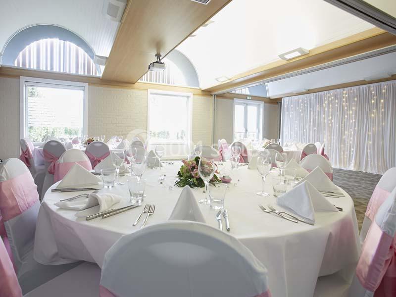 Salle de réception décorée avec des tables rondes, nappes blanches, serviettes pliées et chaises ornées de rubans roses.