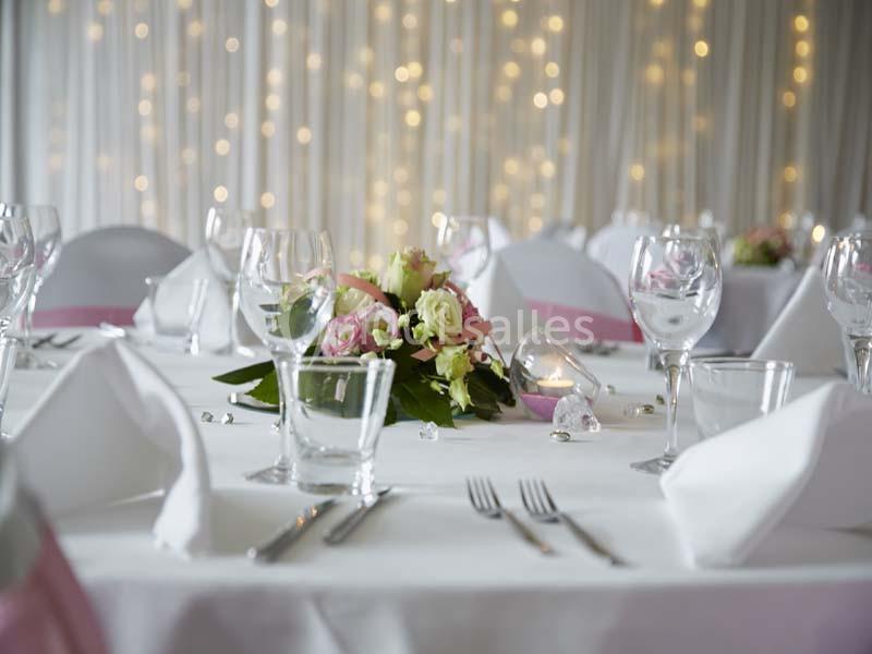 Table décorée pour un événement avec nappes blanches, verres, couverts et un centre de table floral devant un fond lumineux.