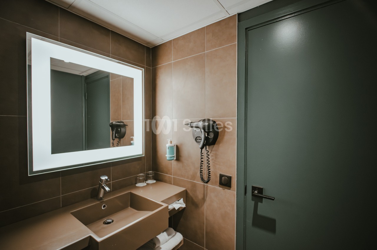 Salle de bain moderne avec lavabo beige, miroir éclairé, sèche-cheveux mural et porte verte.