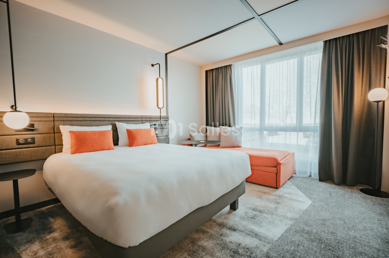 Chambre d'hôtel lumineuse avec un lit double, coussins orange, canapé assorti et grande fenêtre avec rideaux.