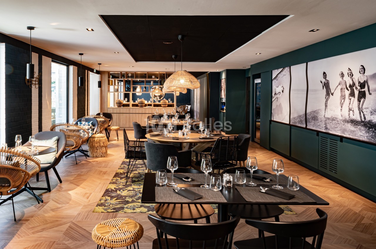 Salle de restaurant moderne avec tables dressées, chaises en rotin et décoration élégante dans des tons sombres et boisés.