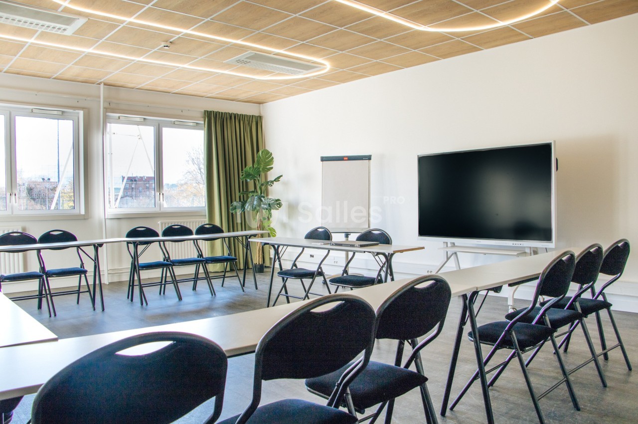 Salle de réunion lumineuse avec tables disposées en U, chaises noires, tableau blanc et écran numérique.