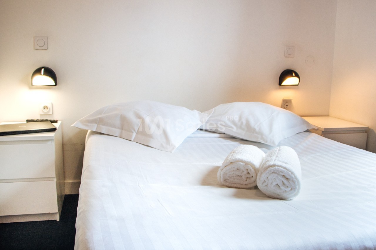 Lit double avec draps blancs, deux oreillers, deux serviettes roulées et lampes murales allumées dans une chambre simple.