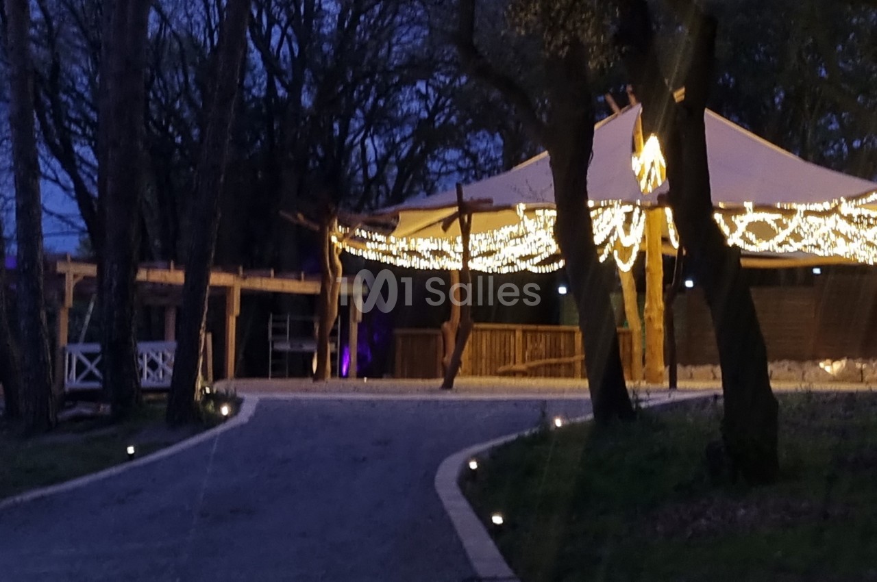 Chemin éclairé menant à une structure en bois couverte, ornée de guirlandes lumineuses, entourée d'arbres.