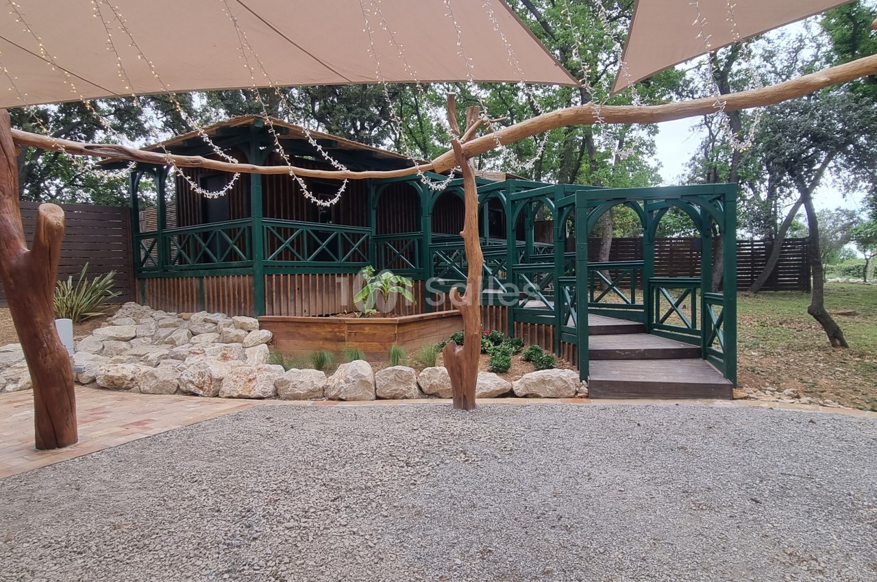 Terrasse en bois avec balustrades vertes, entourée d'arbres et de voiles d'ombrage, sur un sol en gravier et pavés.