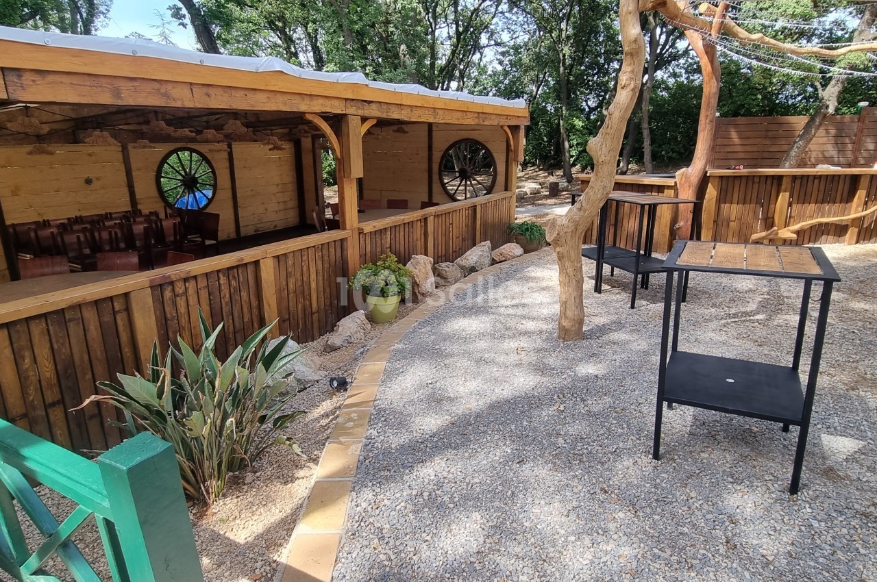 Espace extérieur avec terrasse en bois, tables noires, plantes et arbres, entouré d'une clôture en bois.