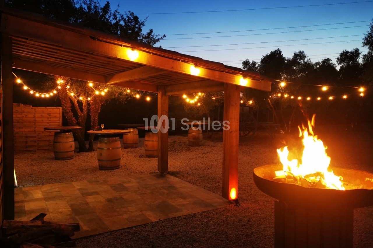 Terrasse extérieure éclairée par des guirlandes lumineuses, avec un brasero allumé et des tonneaux en bois en arrière-plan.