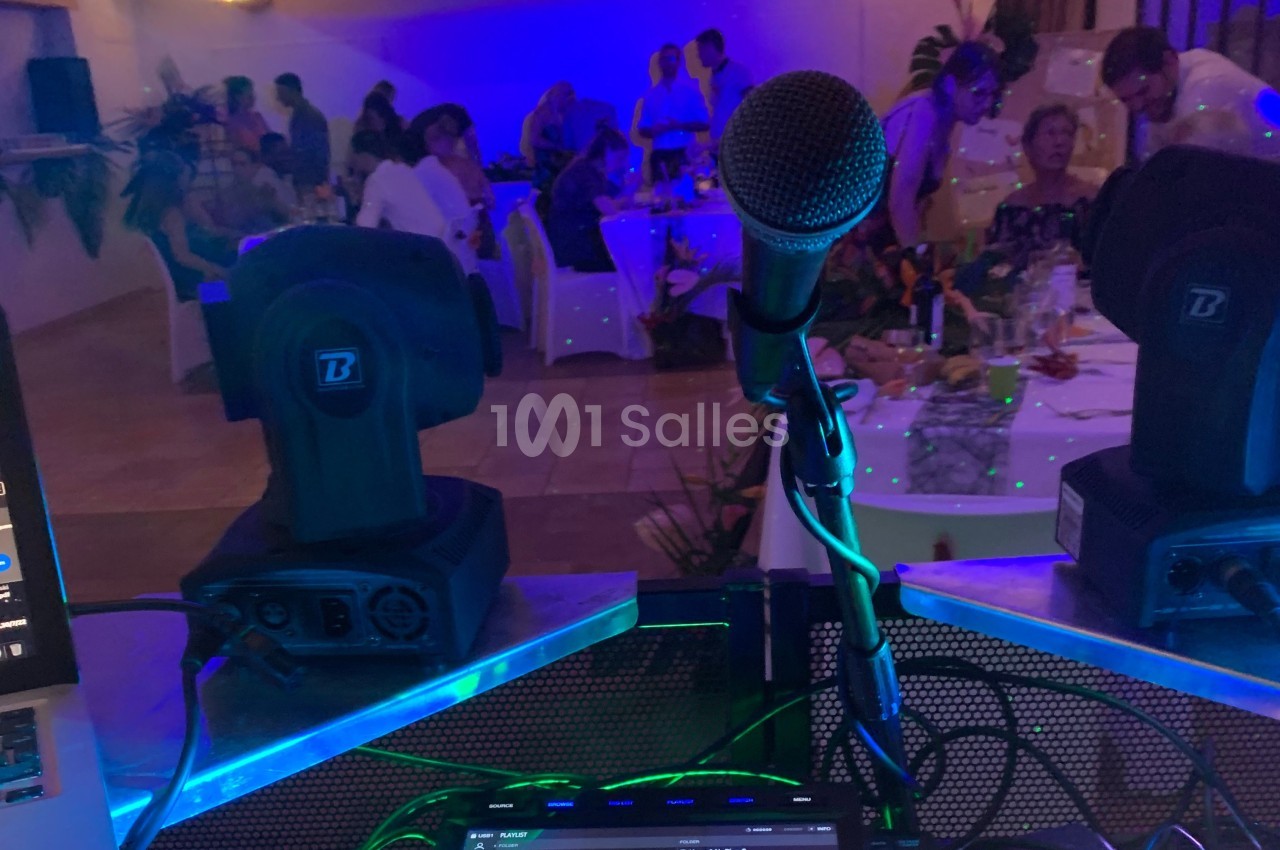 Vue depuis une table de DJ avec un microphone et des projecteurs, face à une salle éclairée en bleu avec des convives.