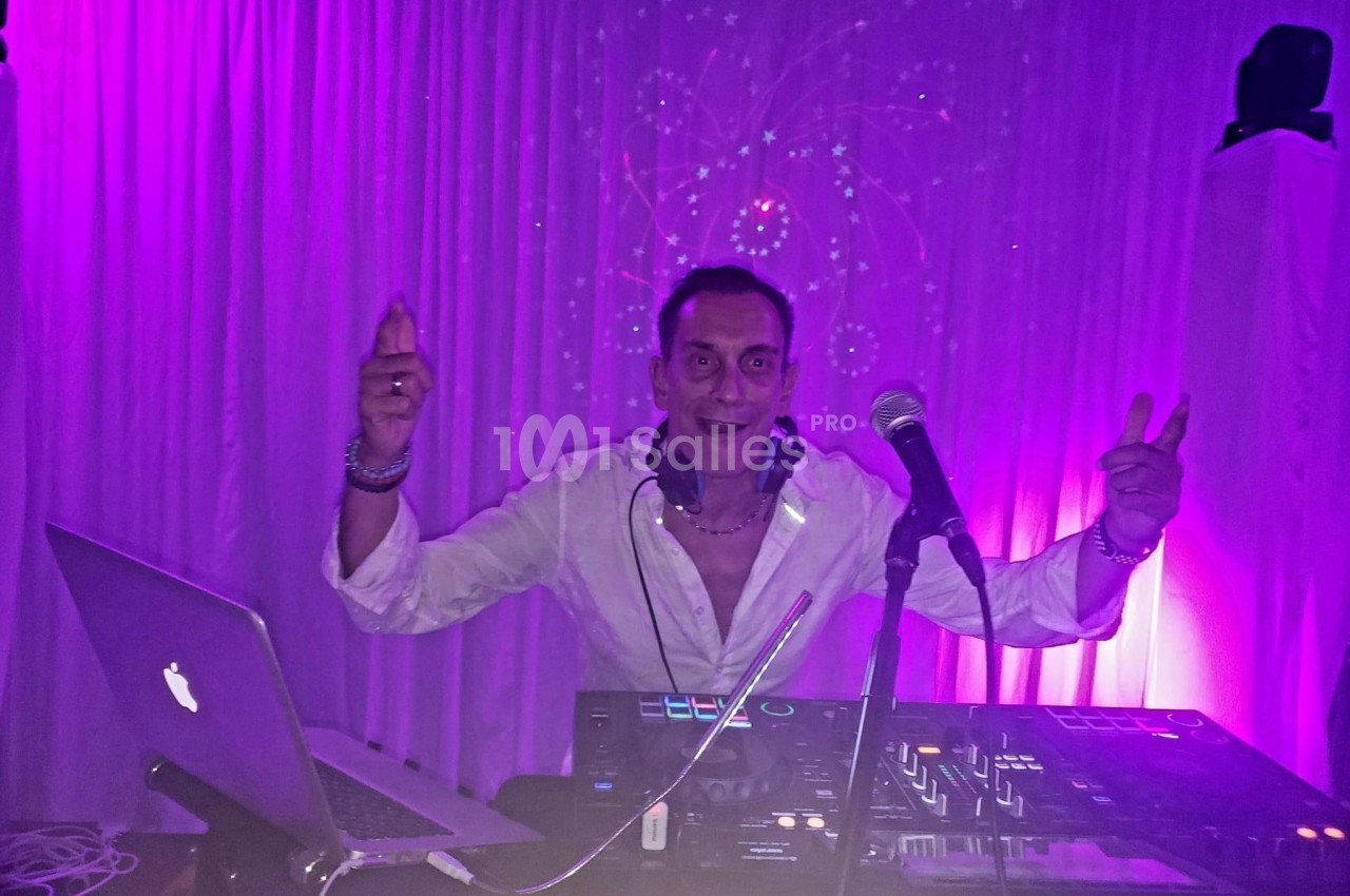 Un DJ en chemise blanche mixe de la musique sur une table de mixage, éclairé par des lumières violettes.