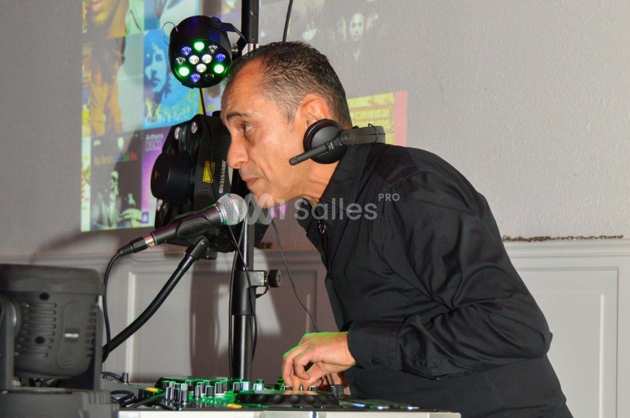 Un DJ en chemise noire mixe de la musique avec un micro et des équipements lumineux en arrière-plan.