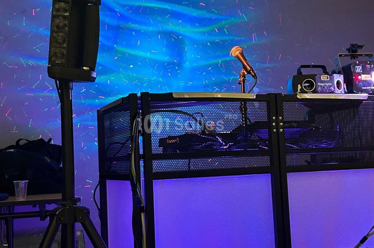 Un pupitre de DJ éclairé avec un micro orange, des équipements audio et des lumières colorées en arrière-plan.