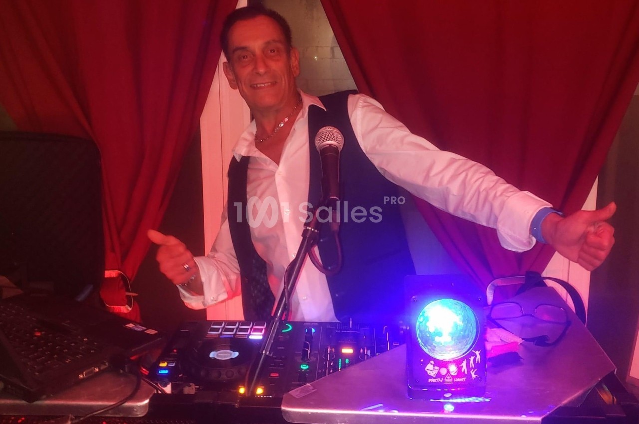 Homme souriant en chemise blanche et gilet noir, debout derrière une table de mixage avec micro et éclairage coloré.