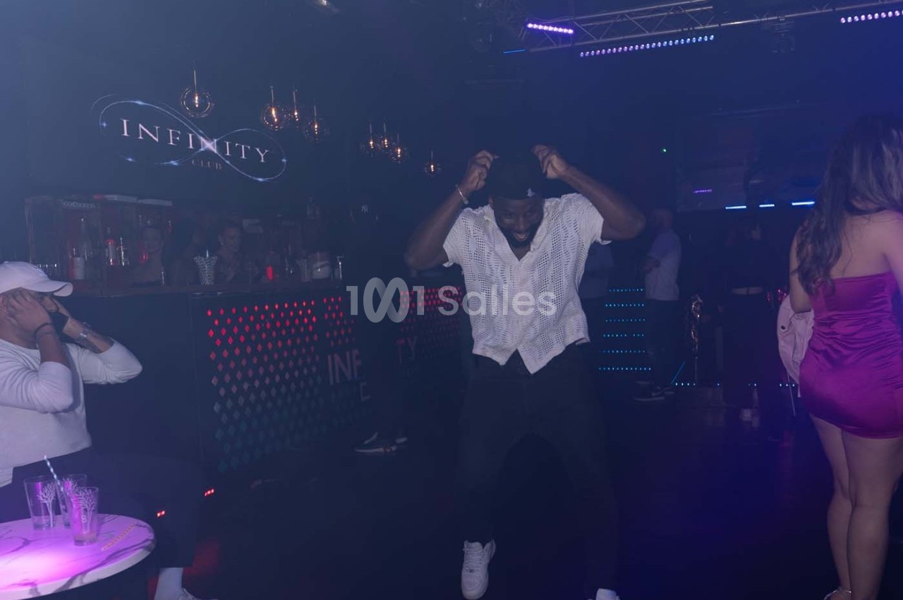 Un homme danse sur une piste de club sombre avec des lumières colorées et un bar en arrière-plan.