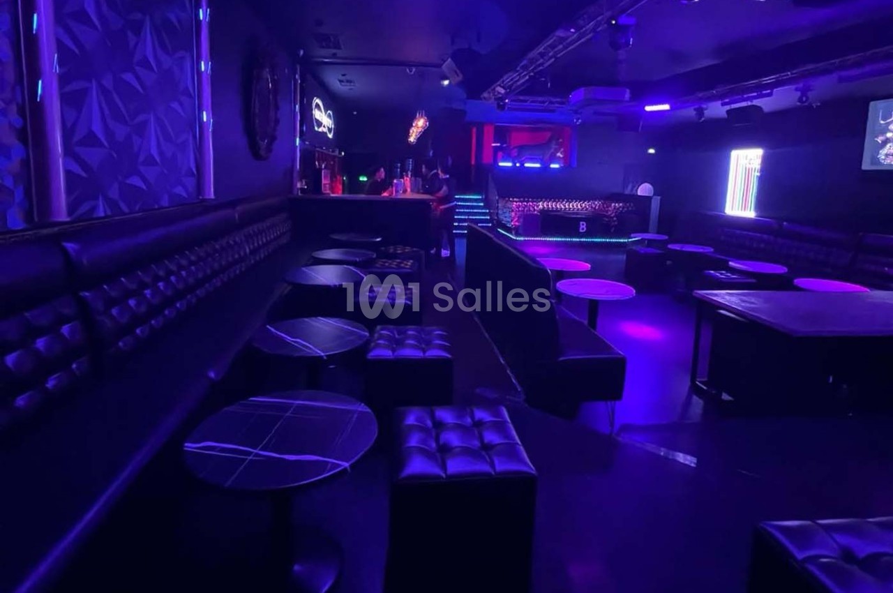 Salle de nuit avec éclairage violet, banquettes noires, tables basses et bar illuminé en arrière-plan.