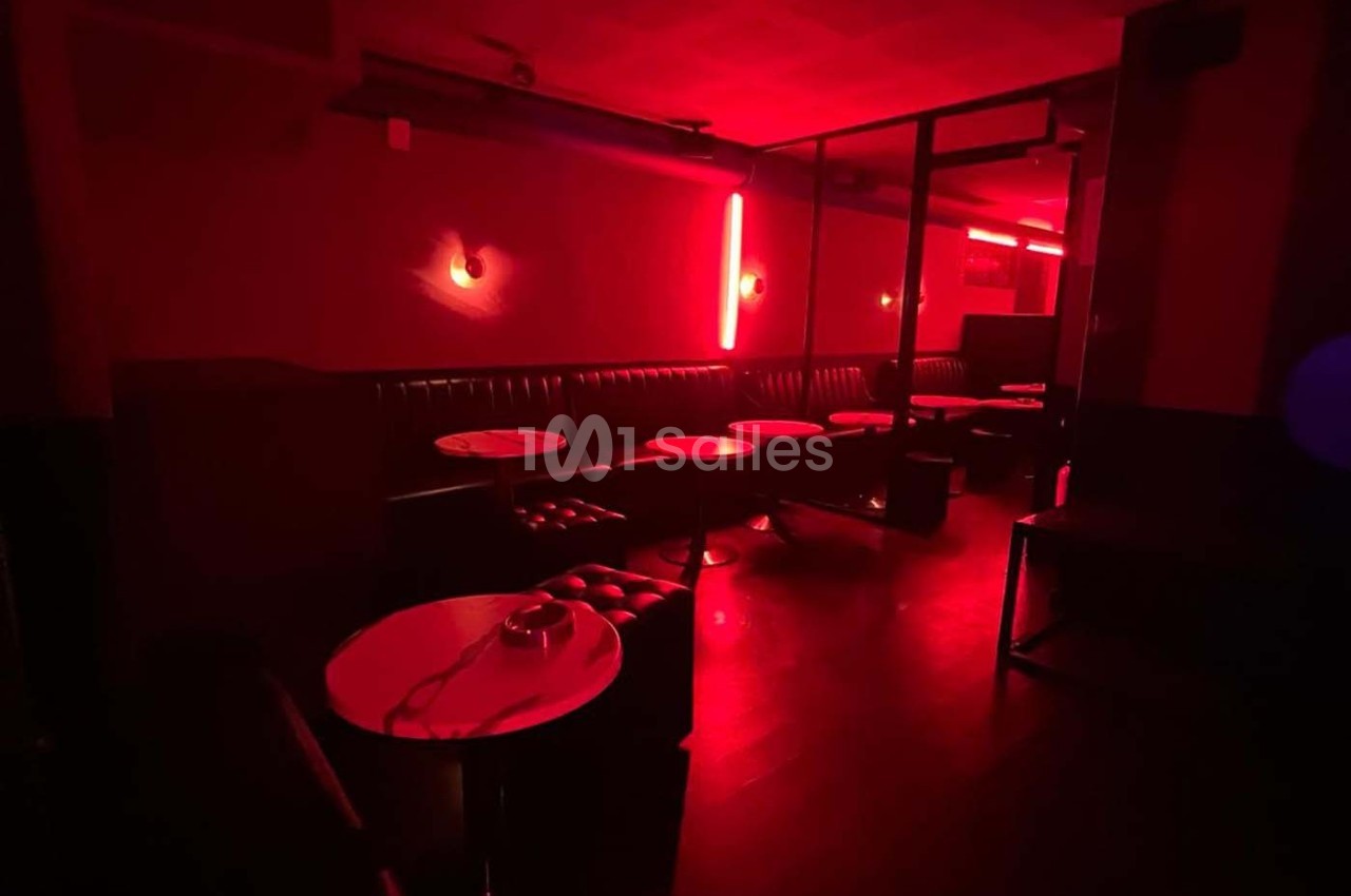 Salle sombre éclairée par des lumières rouges, avec des banquettes, des tables rondes et un sol en bois.