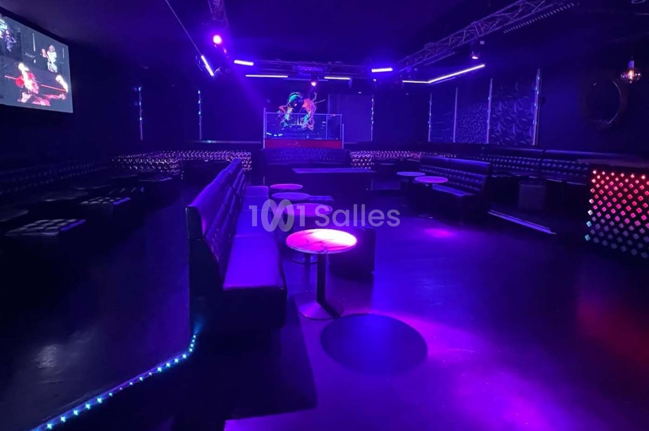 Salle de discothèque vide avec éclairage violet et bleu, banquettes, tables basses et une scène au fond.