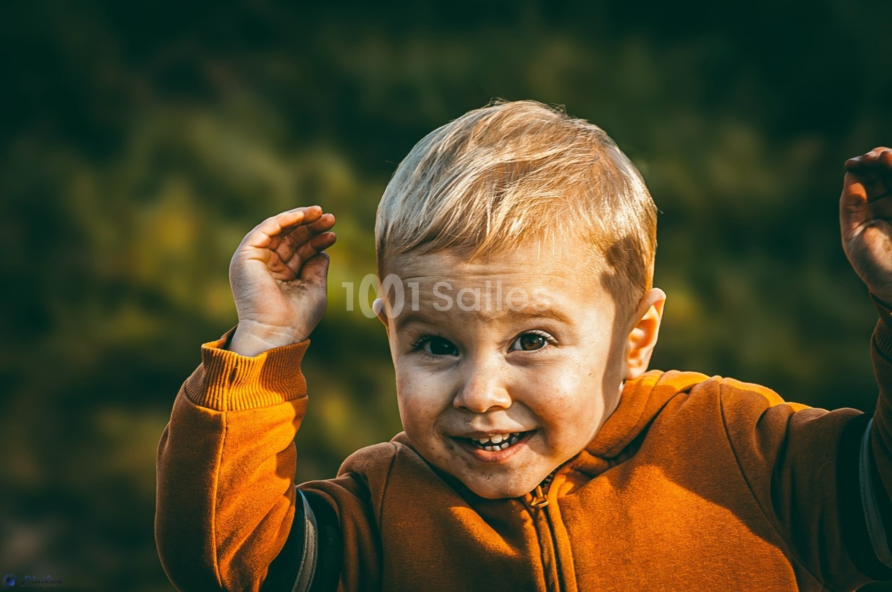 Un enfant blond souriant, vêtu d'un pull orange, levant les mains devant un fond flou de végétation.