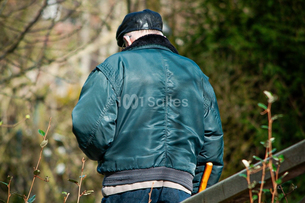 Un homme âgé portant une veste verte et une casquette noire marche avec une canne dans un environnement extérieur.