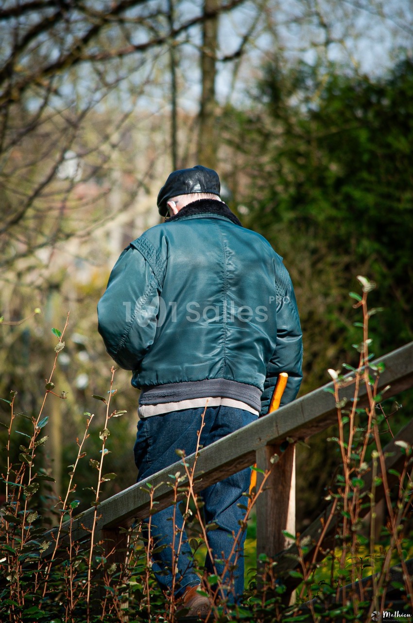 Un homme portant une veste verte et un béret marche sur un sentier boisé, appuyé sur une canne.