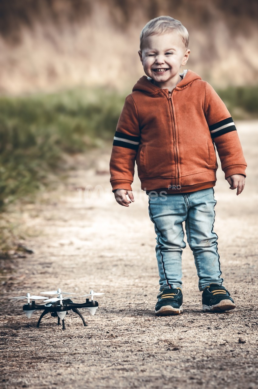 Un enfant souriant en sweat orange regarde un petit drone posé sur un chemin en plein air.