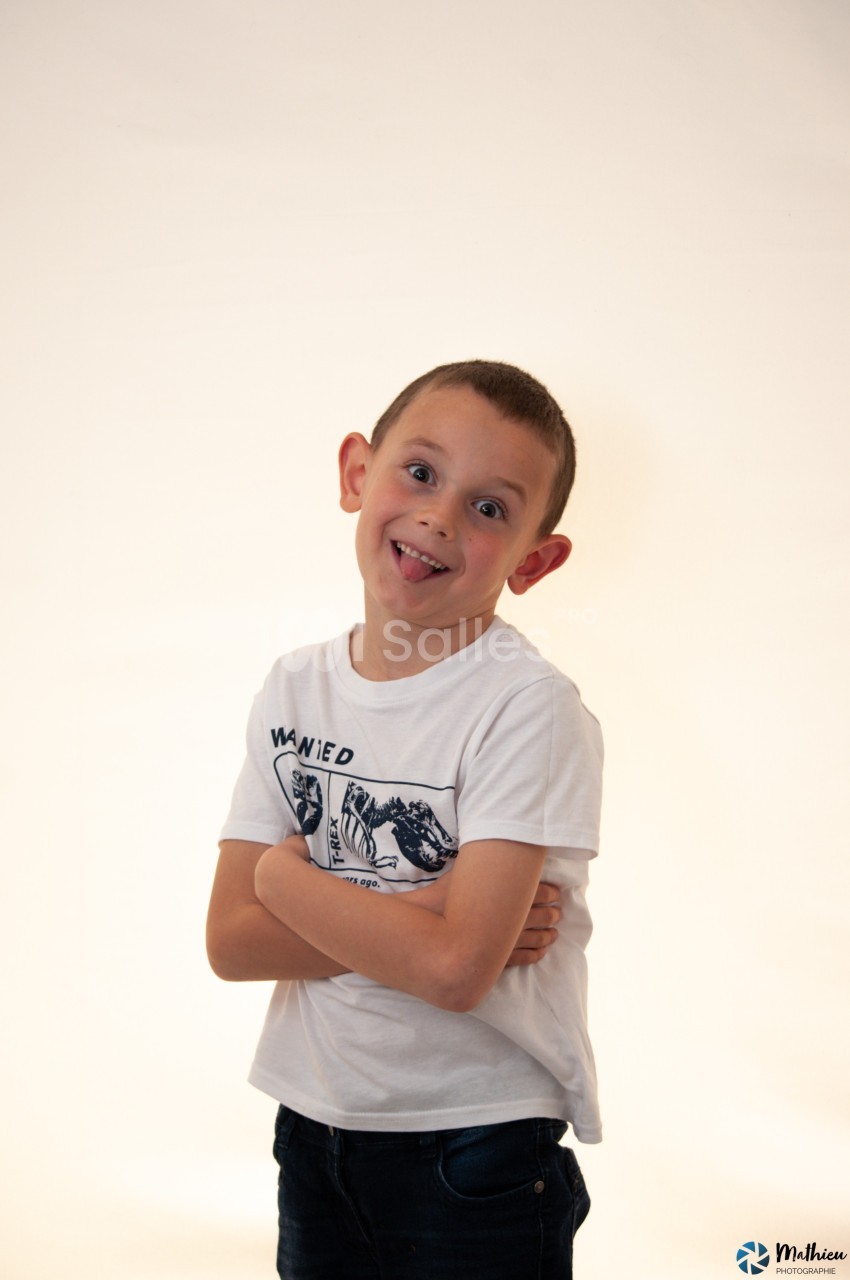Un enfant souriant, bras croisés, tirant la langue, vêtu d'un t-shirt blanc, sur fond clair.