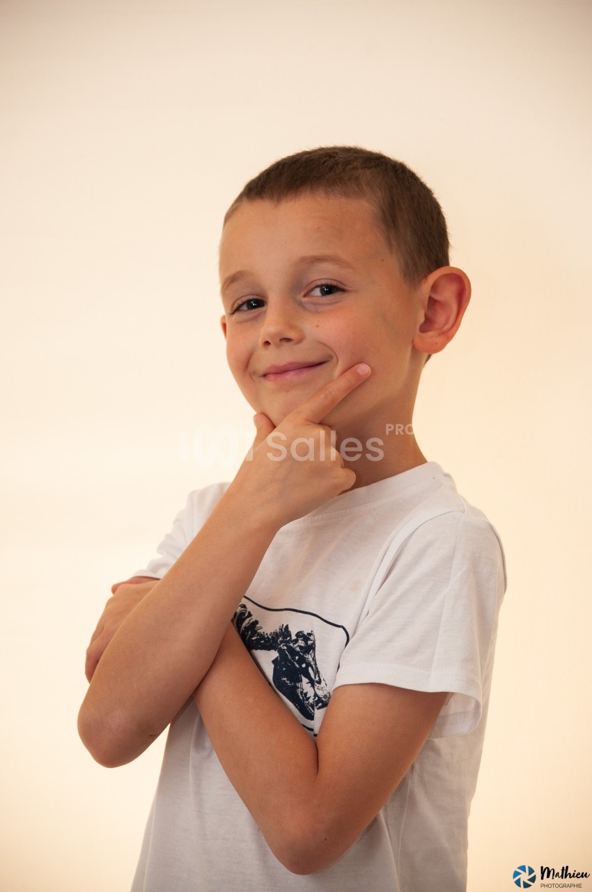 Un enfant souriant en t-shirt blanc pose avec une main sous le menton sur un fond clair.
