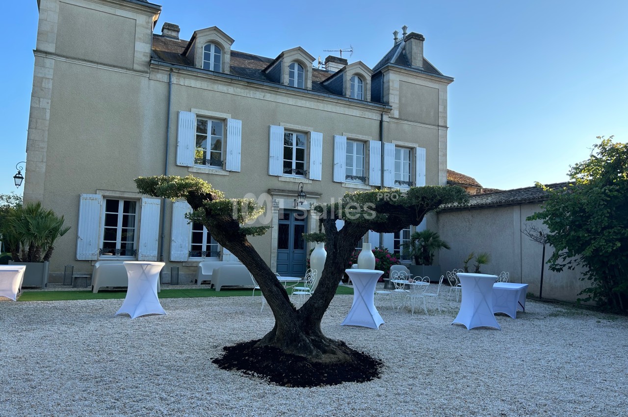 Façade d'une maison en pierre avec un arbre taillé au premier plan, entourée de tables hautes et de chaises blanches.