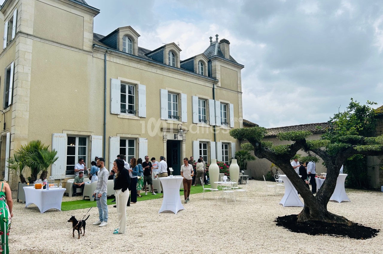 Personnes rassemblées dans la cour d'une grande maison en pierre avec des tables hautes et un arbre taillé au premier plan.