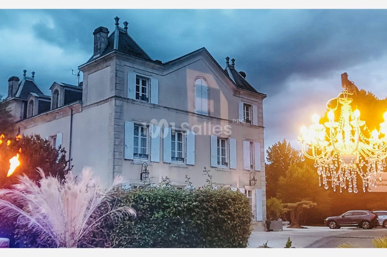 Façade d'un manoir ancien entouré de végétation, avec un lustre illuminé et des torches en premier plan.
