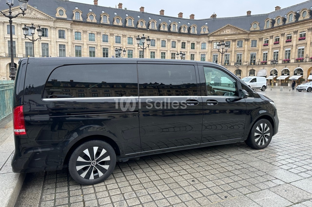 Van noir stationné sur une place pavée devant un bâtiment historique avec des fenêtres alignées et des lampadaires.