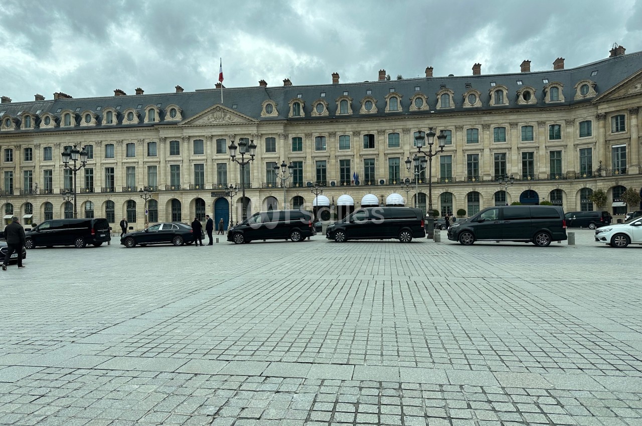 Façade d'un bâtiment historique avec des voitures noires alignées sur une place pavée sous un ciel nuageux.