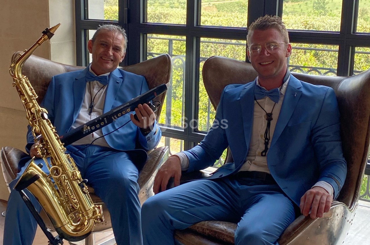 Deux hommes en costume bleu assis sur des fauteuils, l'un tenant un saxophone et l'autre un instrument électronique.