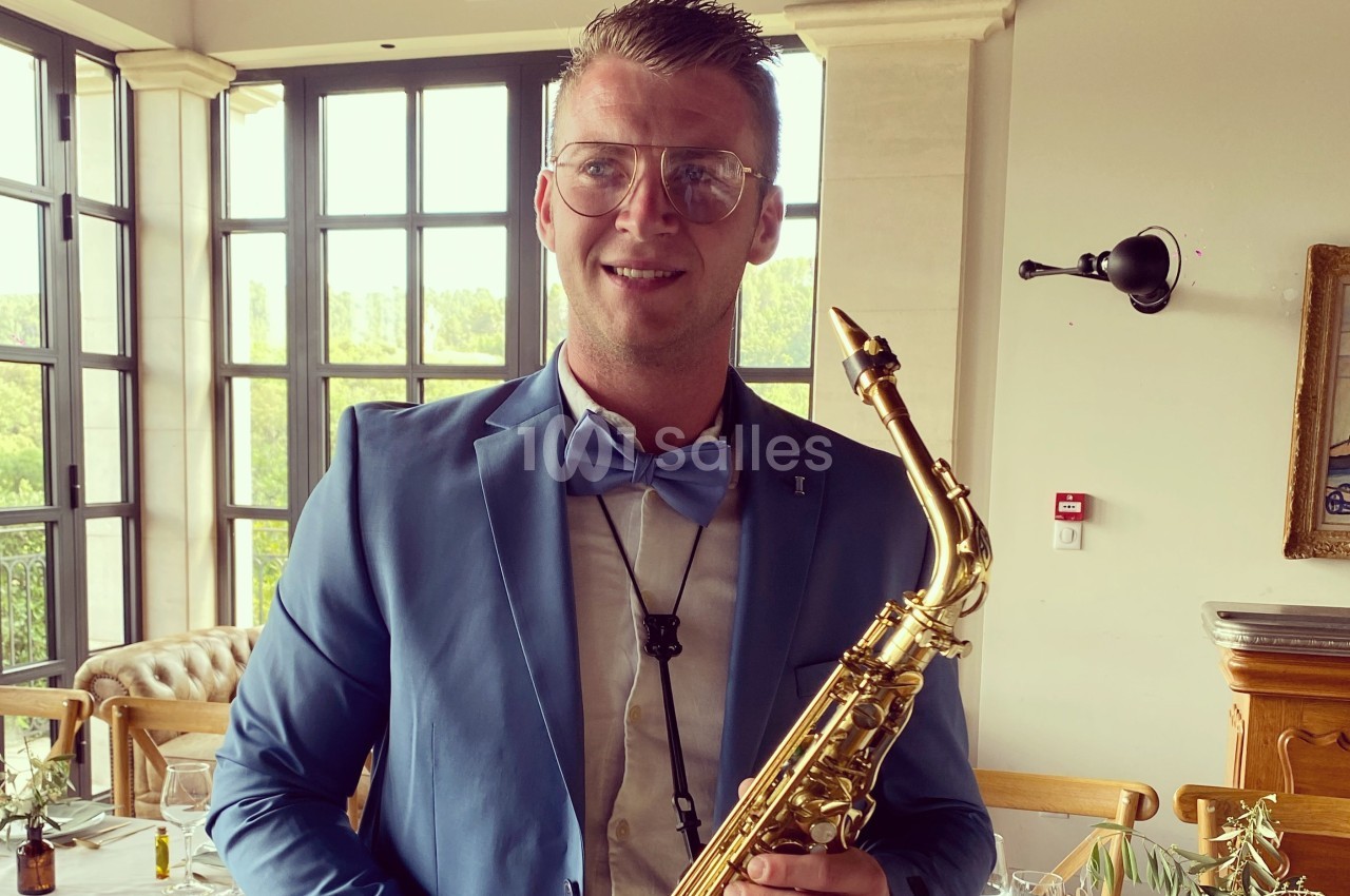 Un homme en costume bleu tient un saxophone dans un intérieur lumineux avec de grandes fenêtres.