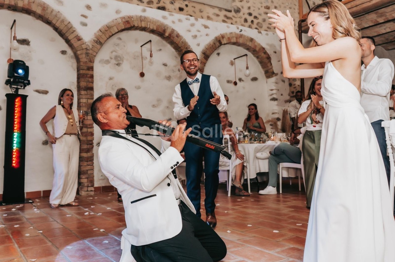 Un musicien en costume blanc joue du saxophone au sol lors d'une fête, entouré d'invités applaudissant.