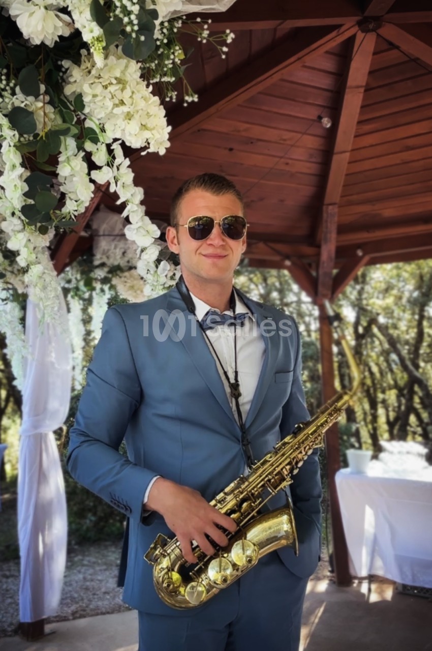 Un homme en costume bleu tient un saxophone sous une pergola décorée de fleurs blanches.