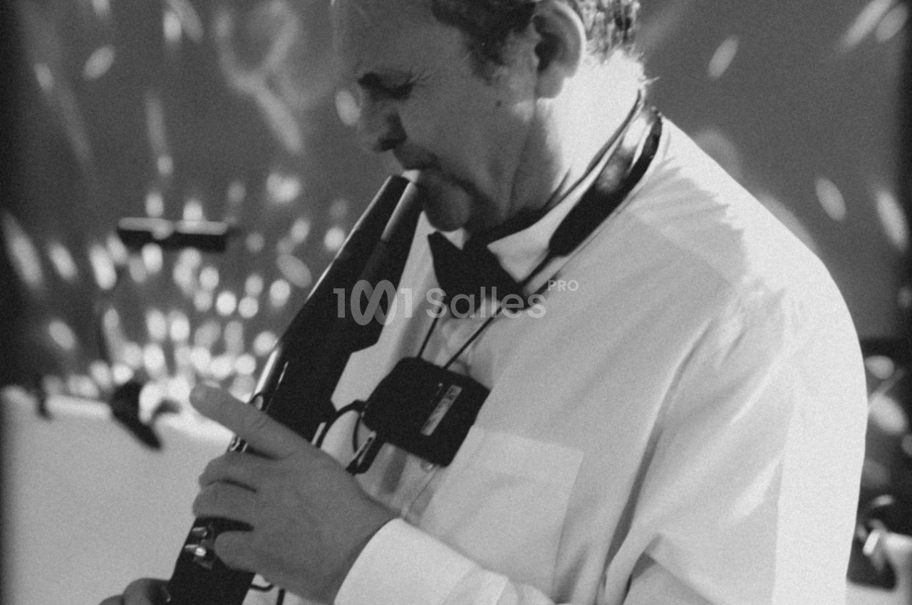 Un musicien en chemise blanche joue d'un instrument à vent électronique dans une ambiance lumineuse tamisée.