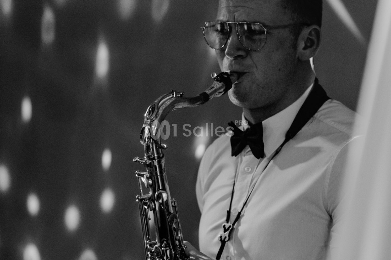 Un musicien en chemise et nœud papillon joue du saxophone dans une ambiance éclairée de lumières tamisées.
