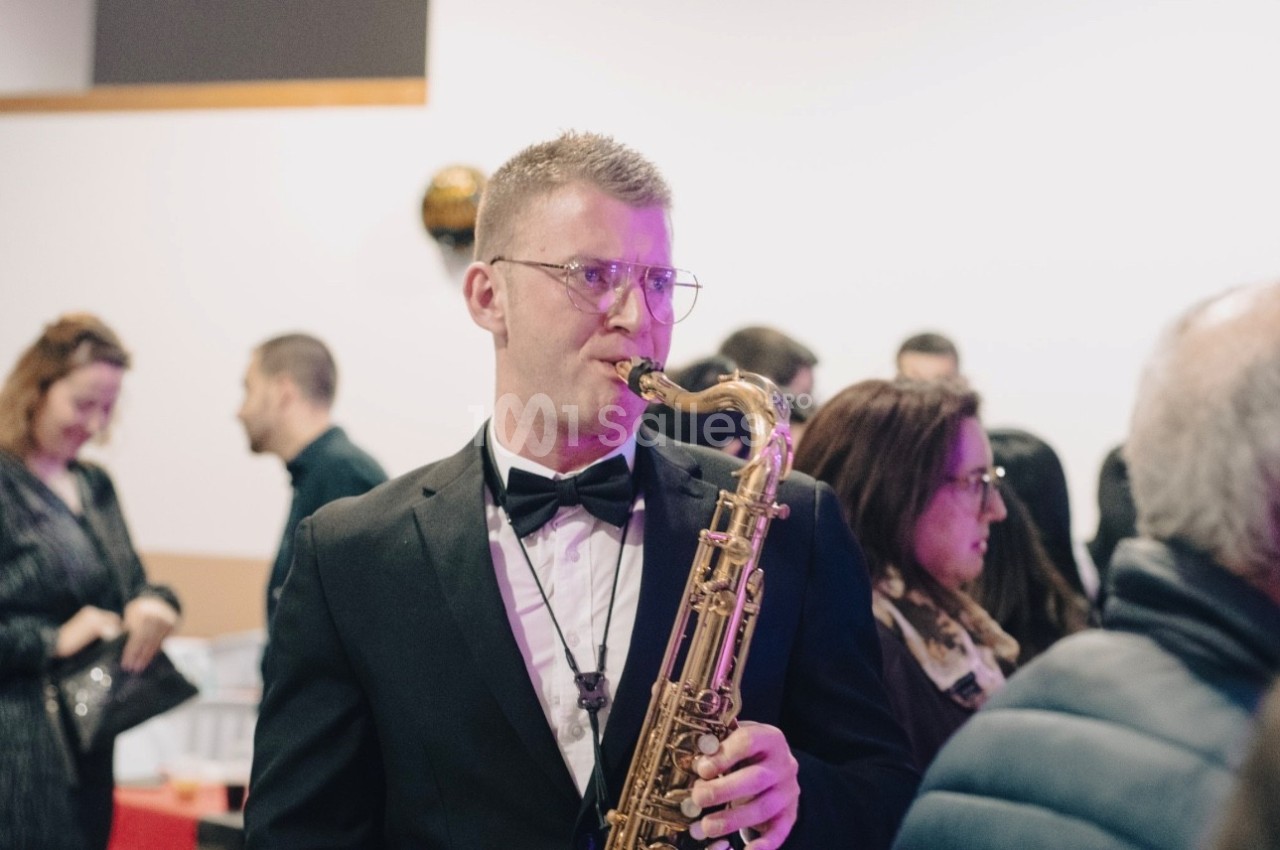 Un musicien en smoking joue du saxophone lors d'un événement avec des personnes en arrière-plan.