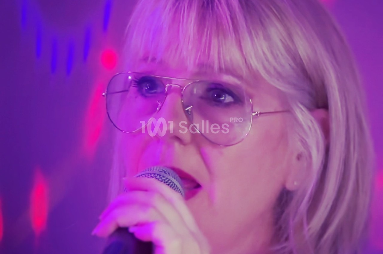 Femme blonde portant des lunettes, chantant au micro sous des lumières violettes et rouges.