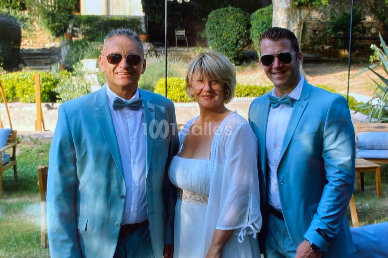 Deux hommes en costume bleu clair et une femme en robe blanche posent ensemble dans un jardin ensoleillé.