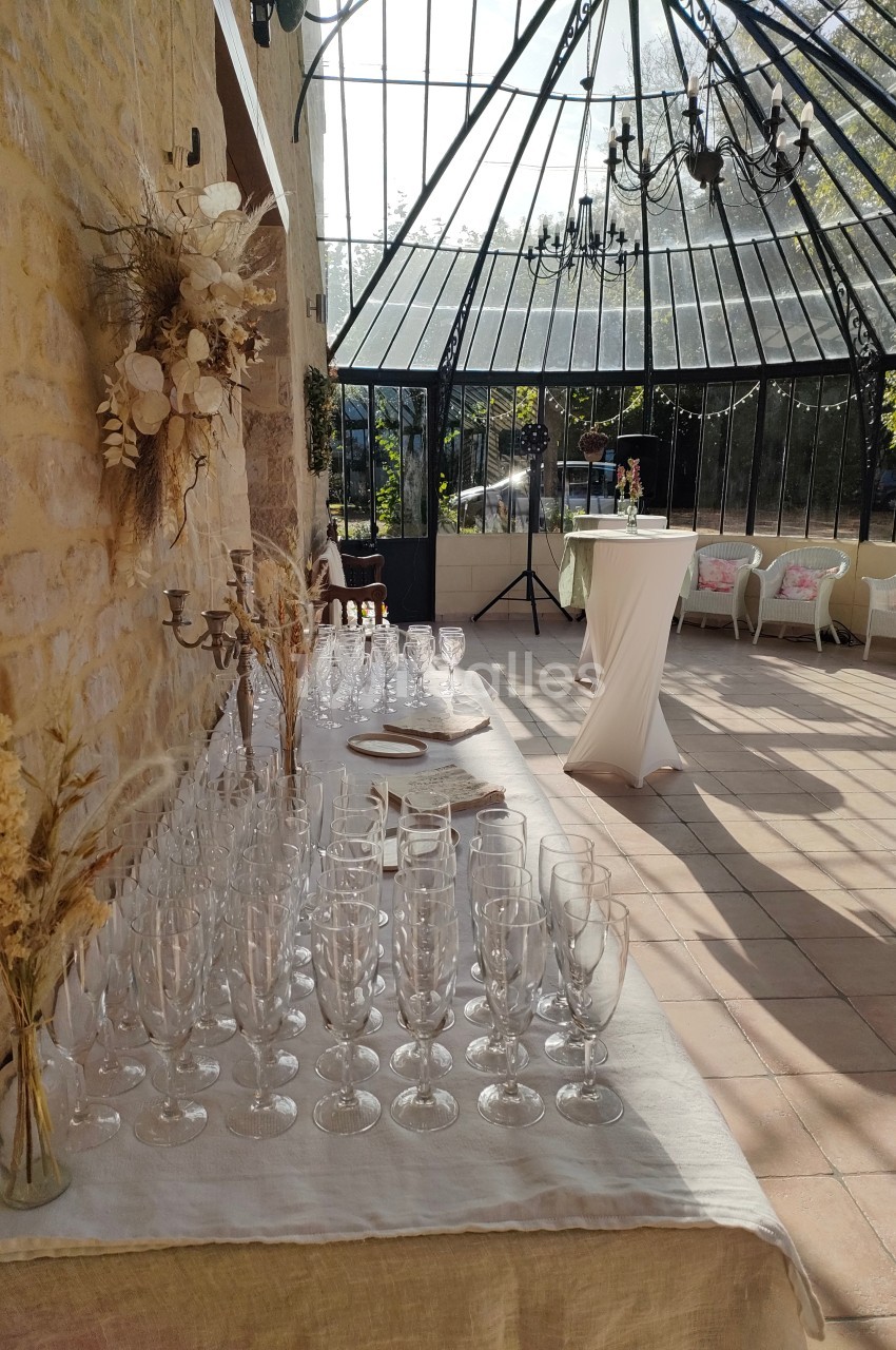 Table dressée avec des verres à pied dans une salle lumineuse avec verrière, décorée de fleurs séchées et mobilier élégant.