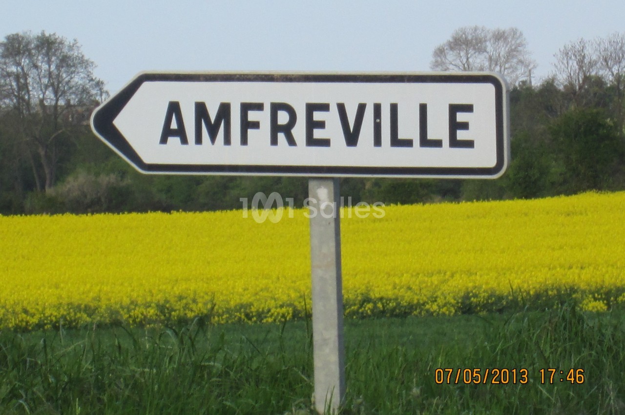Panneau indicateur pointant vers Amfreville, devant un champ de colza et des arbres en arrière-plan.