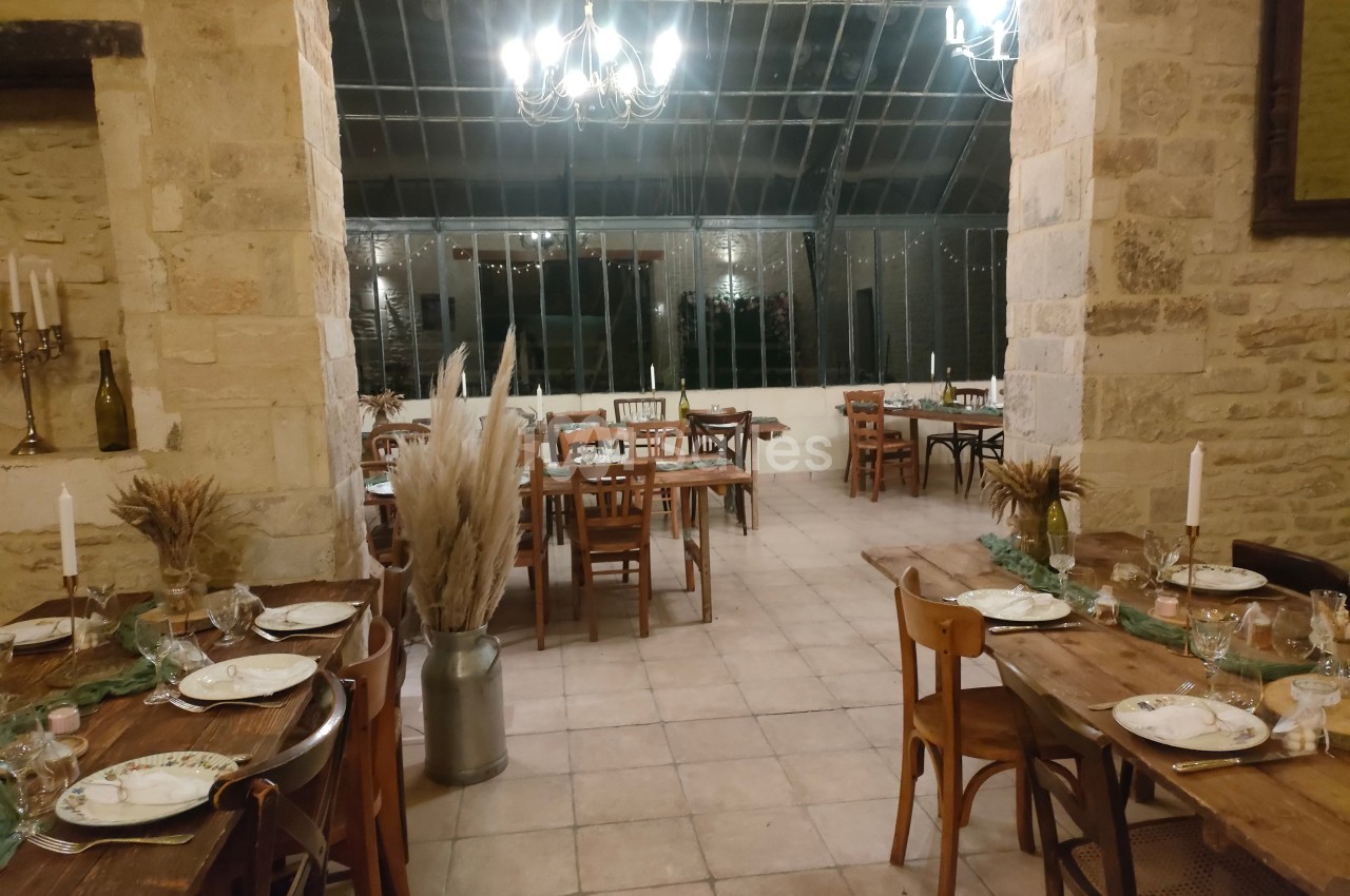 Salle de réception avec tables en bois dressées, décor champêtre et grandes baies vitrées en arrière-plan.