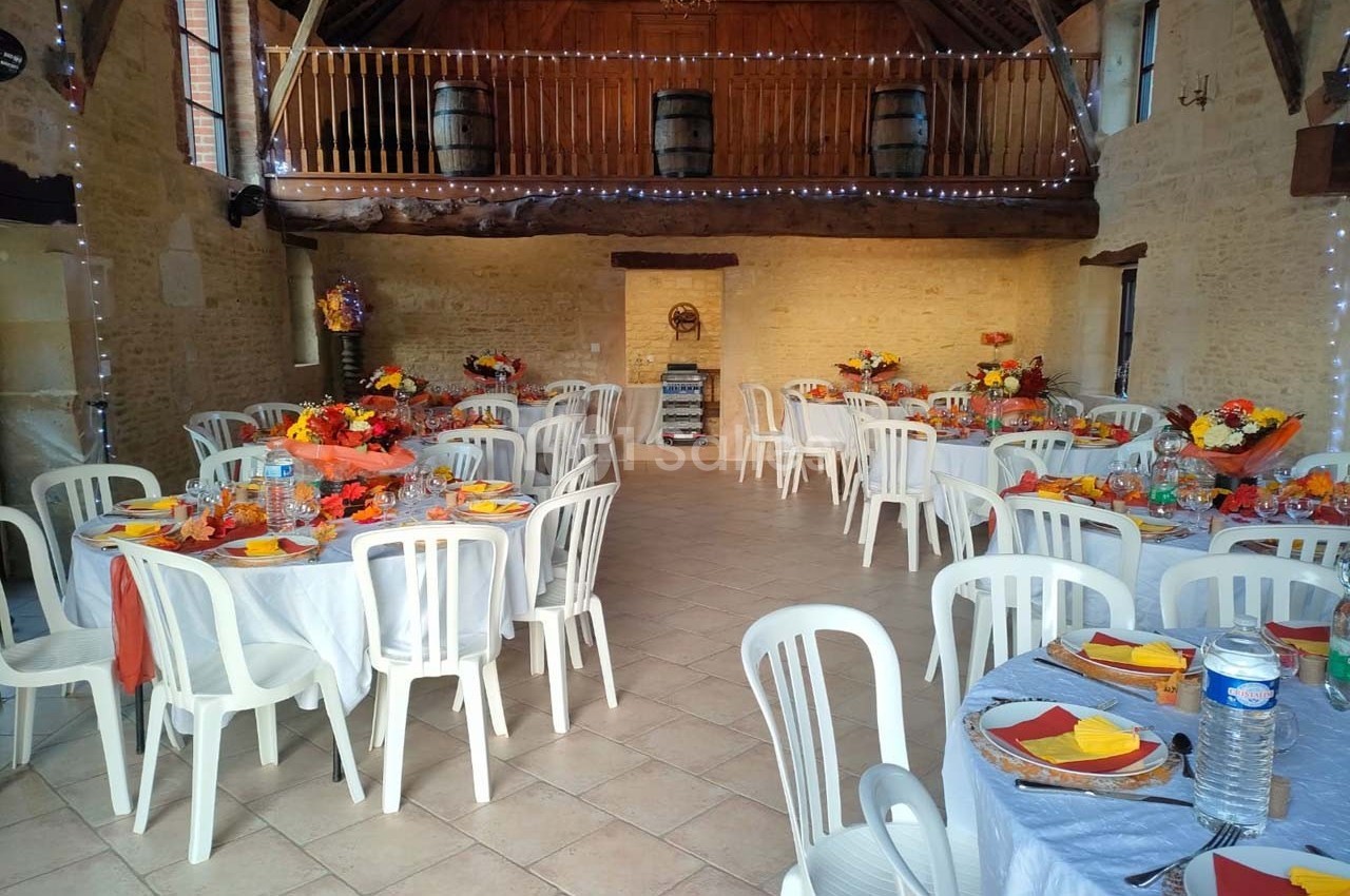 Salle de réception décorée pour un événement, avec tables rondes dressées, chaises blanches et décorations colorées.