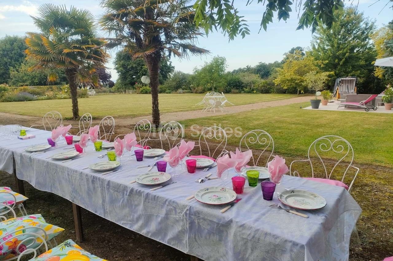 Table dressée en extérieur avec nappes blanches, assiettes, verres colorés et serviettes roses, entourée d'un jardin…